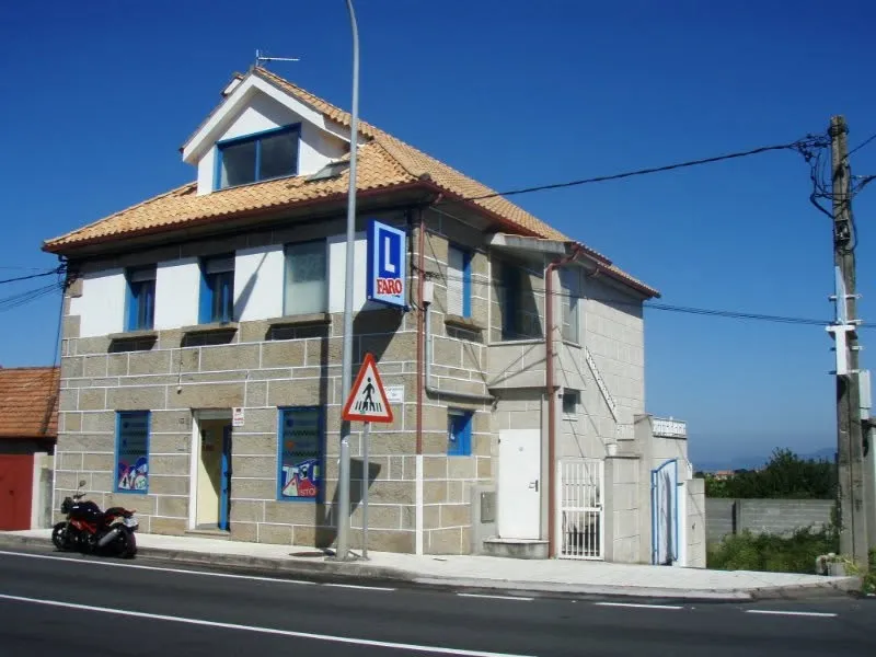 Edificio de piedra de la autoescuela en Coruxo, con el rótulo azul de Faro y una señal de paso de peatones